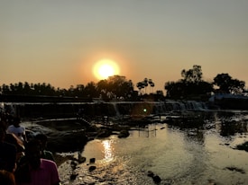 A beautiful sunset over a river with a small waterfall. The silhouette of trees and palm trees are visible against the bright setting sun. There are people standing near the edge of the water, creating a peaceful and serene atmosphere.