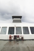 The image shows the exterior side of a large ferry or ship, featuring a series of large windows and a prominent black and white smokestack. The ship's deck is visible, equipped with fire safety equipment including a hose and fire extinguisher. The sky is overcast, creating a gloomy atmosphere.