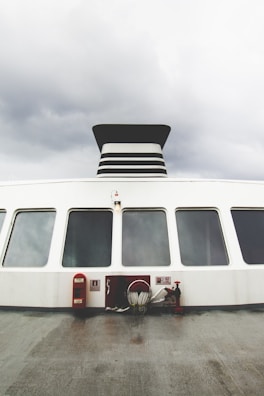 The image shows the exterior side of a large ferry or ship, featuring a series of large windows and a prominent black and white smokestack. The ship's deck is visible, equipped with fire safety equipment including a hose and fire extinguisher. The sky is overcast, creating a gloomy atmosphere.