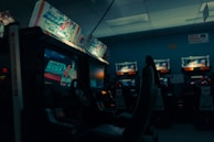 An arcade setting with several racing game machines lined up, featuring illuminated screens displaying a racing game and various promotional artwork above the screens. The ambiance is dimly lit, creating a nostalgic and slightly moody environment.