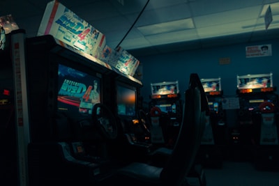 An arcade setting with several racing game machines lined up, featuring illuminated screens displaying a racing game and various promotional artwork above the screens. The ambiance is dimly lit, creating a nostalgic and slightly moody environment.
