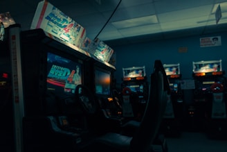 An arcade setting with several racing game machines lined up, featuring illuminated screens displaying a racing game and various promotional artwork above the screens. The ambiance is dimly lit, creating a nostalgic and slightly moody environment.