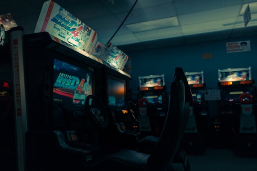 An arcade setting with several racing game machines lined up, featuring illuminated screens displaying a racing game and various promotional artwork above the screens. The ambiance is dimly lit, creating a nostalgic and slightly moody environment.