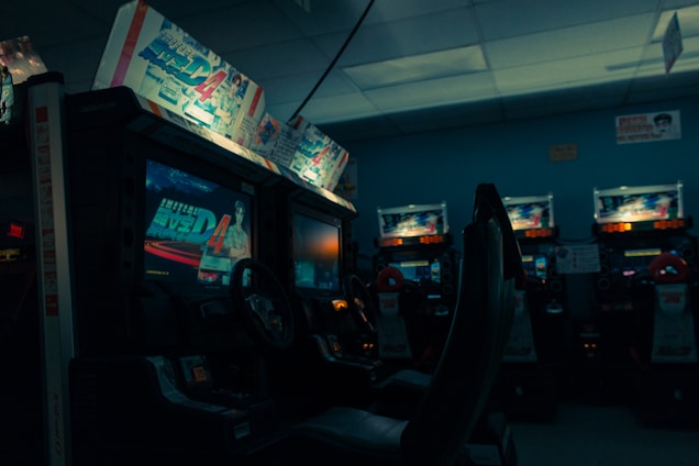 An arcade setting with several racing game machines lined up, featuring illuminated screens displaying a racing game and various promotional artwork above the screens. The ambiance is dimly lit, creating a nostalgic and slightly moody environment.