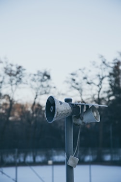 A weathered, old loudspeaker mounted on a tall pole with a blurred background of bare trees and a fence. The sky is clear and blue, indicating a calm and cold day. The speakers appear to be positioned outdoors in a somewhat isolated area.