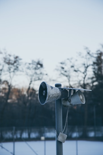 A weathered, old loudspeaker mounted on a tall pole with a blurred background of bare trees and a fence. The sky is clear and blue, indicating a calm and cold day. The speakers appear to be positioned outdoors in a somewhat isolated area.