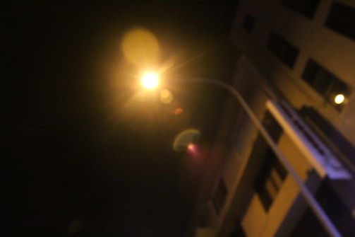 A bright, glowing streetlight against a dark night sky, with nearby buildings partially visible. The light creates lens flares and illuminates parts of the scene.