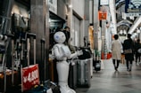 A humanoid robot stands in an indoor shopping arcade next to a display of suitcases. People are walking through the arcade, which is lined with various shops. The area is well-lit, with a mix of natural and artificial lighting. Signs and advertisements in Japanese can be seen around, indicating the prices of products.