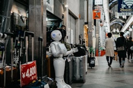 A humanoid robot stands in an indoor shopping arcade next to a display of suitcases. People are walking through the arcade, which is lined with various shops. The area is well-lit, with a mix of natural and artificial lighting. Signs and advertisements in Japanese can be seen around, indicating the prices of products.