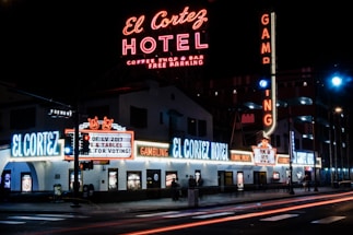 A brightly lit street corner featuring the El Cortez Hotel with numerous neon signs advertising various amenities such as a coffee shop, bar, and gambling. The hotel name is prominently displayed in large neon letters, and there are additional signs including 'Gambling' and 'Free Parking'. People can be seen walking near the hotel's entrance.