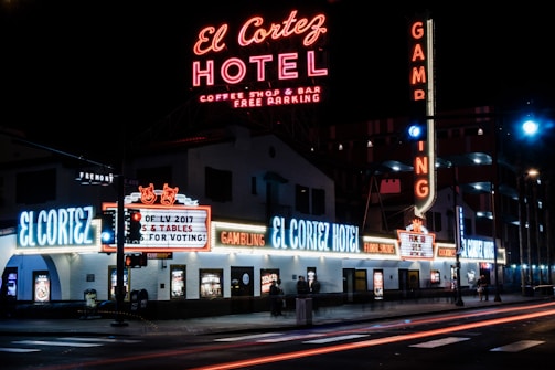 A brightly lit street corner featuring the El Cortez Hotel with numerous neon signs advertising various amenities such as a coffee shop, bar, and gambling. The hotel name is prominently displayed in large neon letters, and there are additional signs including 'Gambling' and 'Free Parking'. People can be seen walking near the hotel's entrance.
