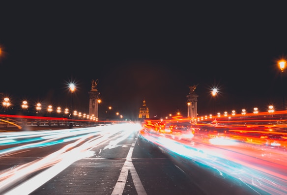 A long exposure photograph of a bustling city street at night, showing light trails from moving vehicles. The background features ornate lampposts and two large sculptures flanking a central building with a dome, possibly a historic monument, all lit up against a dark sky.