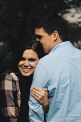 A couple stands closely together with the woman smiling warmly. She has long dark hair and is wearing a plaid jacket. Her hand is gently resting on the man's shoulder, displaying an engagement ring. The man is dressed in a light blue shirt and is turned away, nuzzling affectionately against her head.
