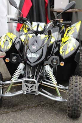 A black and yellow quad bike is parked, featuring a design with bold graphics on the body. The front suspension and tires are clearly visible, highlighting the rugged build. The background includes some red and black fabric, adding a backdrop to the image.