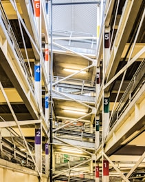 The image shows an interior view of a multi-level parking garage. Multiple floors are visible, connected by ramps marked with colorful numbers corresponding to different levels. The structure is composed of steel beams and concrete, with fluorescent lighting illuminating the space.
