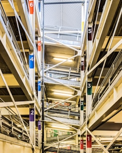 The image shows an interior view of a multi-level parking garage. Multiple floors are visible, connected by ramps marked with colorful numbers corresponding to different levels. The structure is composed of steel beams and concrete, with fluorescent lighting illuminating the space.