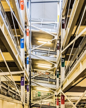 The image shows an interior view of a multi-level parking garage. Multiple floors are visible, connected by ramps marked with colorful numbers corresponding to different levels. The structure is composed of steel beams and concrete, with fluorescent lighting illuminating the space.