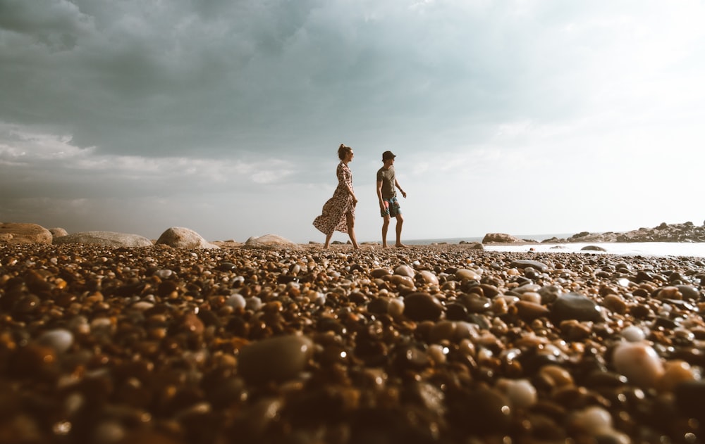 man and woman walking on stones near beach under blue sky