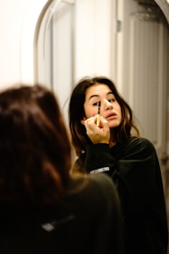 A smiling woman applying eyebrow makeup in front of a mirror with soft lighting.