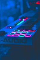 A keyboard synthesizer illuminated by vibrant neon lights, highlighting its keys and control pads in shades of blue, pink, and green. The scene has a futuristic and electronic ambiance.