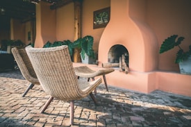 Two wicker chairs are positioned on a cobblestone patio in front of a unique fireplace with an earthy orange plaster finish. The area is surrounded by large green potted plants, creating a serene and natural atmosphere. The sunlight casts soft shadows over the scene, enhancing the warm and inviting mood.