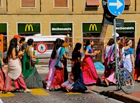 A group of people wearing colorful traditional attire walk along a city street. Behind them, two McDonald's signs are visible against a building, and a bicycle is parked near a traffic sign.
