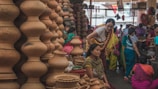 A bustling market scene where several women are gathered around a variety of clay pots and earthenware. The pots are stacked in unique shapes, displaying a rich cultural craft. The women are dressed in colorful traditional attire, engaging in conversation or examining the pottery.