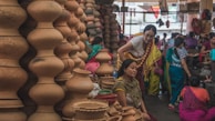 A bustling market scene where several women are gathered around a variety of clay pots and earthenware. The pots are stacked in unique shapes, displaying a rich cultural craft. The women are dressed in colorful traditional attire, engaging in conversation or examining the pottery.