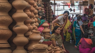 A bustling market scene where several women are gathered around a variety of clay pots and earthenware. The pots are stacked in unique shapes, displaying a rich cultural craft. The women are dressed in colorful traditional attire, engaging in conversation or examining the pottery.