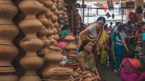 A bustling market scene where several women are gathered around a variety of clay pots and earthenware. The pots are stacked in unique shapes, displaying a rich cultural craft. The women are dressed in colorful traditional attire, engaging in conversation or examining the pottery.