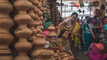 A bustling market scene where several women are gathered around a variety of clay pots and earthenware. The pots are stacked in unique shapes, displaying a rich cultural craft. The women are dressed in colorful traditional attire, engaging in conversation or examining the pottery.