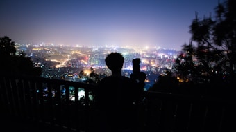 A silhouette of a person holding a camera overlooks a vibrant, illuminated cityscape at night. The view is expansive, showing countless twinkling lights and multicolored buildings stretching into the distance. Trees partially frame the scene, adding depth to the foreground.