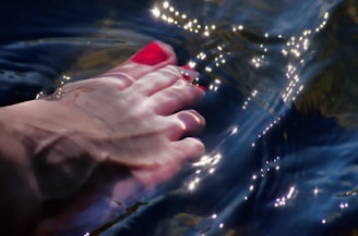 A close-up of a human foot with toes painted red is partially submerged in water. The sunlight creates shimmering reflections and ripples on the water surface.