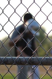 A chain-link fence is in sharp focus in the foreground, with a blurred figure in the background. The setting appears to be an outdoor sports field, indicated by the uniform and helmet worn by the figure.