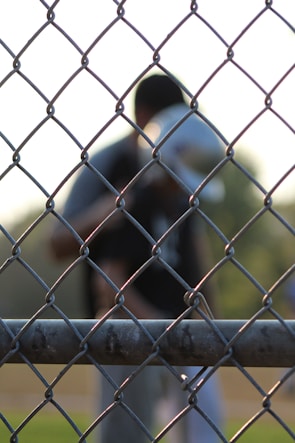 A chain-link fence is in sharp focus in the foreground, with a blurred figure in the background. The setting appears to be an outdoor sports field, indicated by the uniform and helmet worn by the figure.