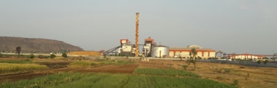 A large industrial complex situated in a semi-arid landscape with patches of green fields in the foreground. The facility includes multiple structures such as silos, large cylindrical tanks, and a prominent chimney. The horizon features a low mountain range under a clear sky.
