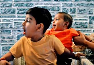 Two children are positioned next to a textured stone or brick wall. The older child, wearing a tan shirt, has an expression of surprise or excitement. The younger child, held by an adult's hands, is also looking in the same direction with an animated expression while wearing a bright orange outfit.