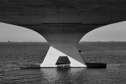 A striking black and white photograph of a large concrete bridge stretching across a wide body of water. The bridge features a geometric support structure with a prominent triangular form. In the background, distant wind turbines are faintly visible on the horizon. The scene is serene and minimalistic.