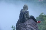A person wearing a hooded jacket and a backpack sits on a rocky ledge, overlooking a misty landscape. The individual is facing away, creating a sense of solitude and reflection amidst the natural surroundings.