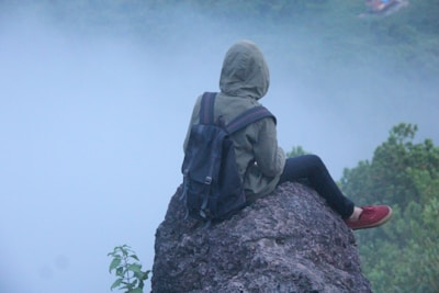 A person wearing a hooded jacket and a backpack sits on a rocky ledge, overlooking a misty landscape. The individual is facing away, creating a sense of solitude and reflection amidst the natural surroundings.