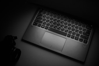 A grayscale image of a laptop keyboard and touchpad is prominently displayed. The lighting creates a dramatic, high-contrast effect, emphasizing the keys and the sleek design of the laptop. In the dim lighting, there is a hint of a camera lens partially visible in the shadowy foreground, suggesting a photography-related theme.