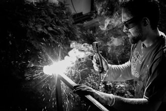 A person is engaged in welding, holding a welding torch that emits bright sparks and smoke. The scene is set outdoors with foliage in the background, and the person is wearing safety glasses and a casual shirt.