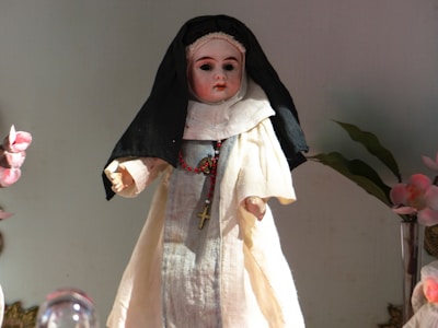 A doll dressed in a nun's habit, featuring a black veil and a white robe, adorned with a cross necklace. The doll is surrounded by decorative elements, including flowers and possibly other objects.