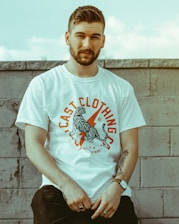 A person with a beard is leaning against a concrete wall wearing a graphic tee with a bold design featuring a tiger and text that reads 'Cast Clothing'. The background suggests an outdoor setting with muted tones.