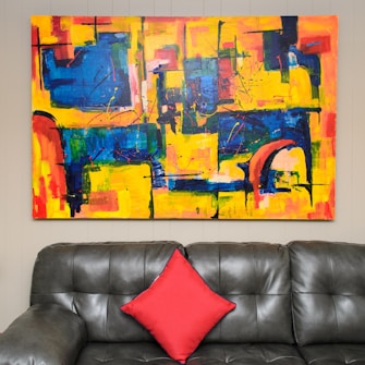 An abstract painting with bold colors hangs on a wall above a dark gray leather sofa. The artwork features geometric patterns with predominant colors of yellow, blue, and red. A single red cushion is placed in the center of the sofa.