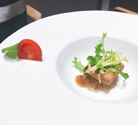A white plate with a gourmet dish. The center features a small portion of meat topped with crispy bread and fresh green salad leaves. A light brown sauce complements the dish. On the edge of the plate, a quarter of a cherry tomato and a sprig of green beans are arranged decoratively.