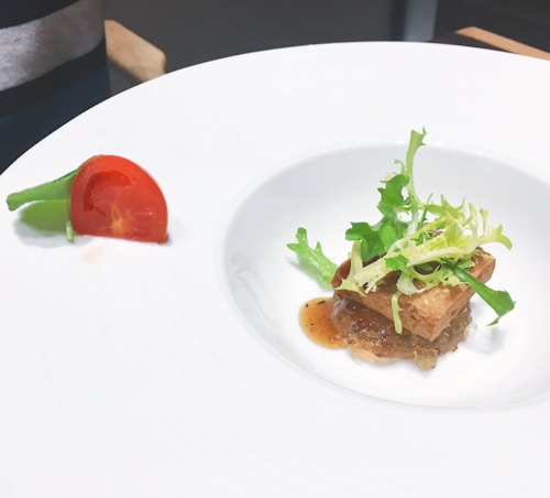 A white plate with a gourmet dish. The center features a small portion of meat topped with crispy bread and fresh green salad leaves. A light brown sauce complements the dish. On the edge of the plate, a quarter of a cherry tomato and a sprig of green beans are arranged decoratively.