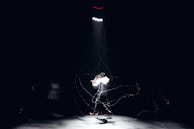 A person standing alone under a bright spotlight in a dark environment. The figure appears to be engaging in a dynamic motion with motion blur effects, creating a sense of movement. There are faint lines and particles surrounding the person, adding an ethereal quality to the scene.