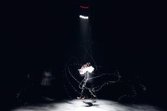 A person standing alone under a bright spotlight in a dark environment. The figure appears to be engaging in a dynamic motion with motion blur effects, creating a sense of movement. There are faint lines and particles surrounding the person, adding an ethereal quality to the scene.