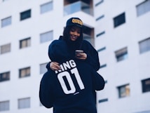 A person wearing a black hoodie with 'KING 01' written on the back carries someone else on their shoulders. The person being carried is wearing a cap with the word 'QUEEN' and is holding a smartphone. They are against the backdrop of a modern building with windows.